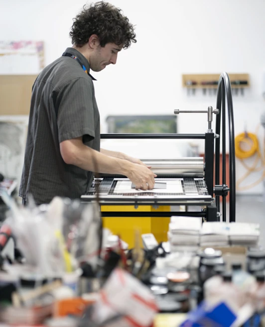Person operating a large printmaking press in an art studio with various supplies in the foreground. Person operating a large printmaking press in an art studio with various supplies in the foreground.